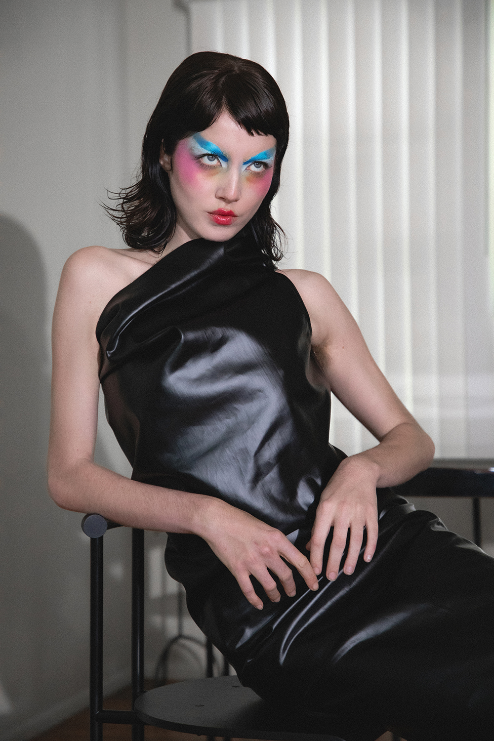 Autry seated in a black leather dress with colorful makeup looking intently past the camera